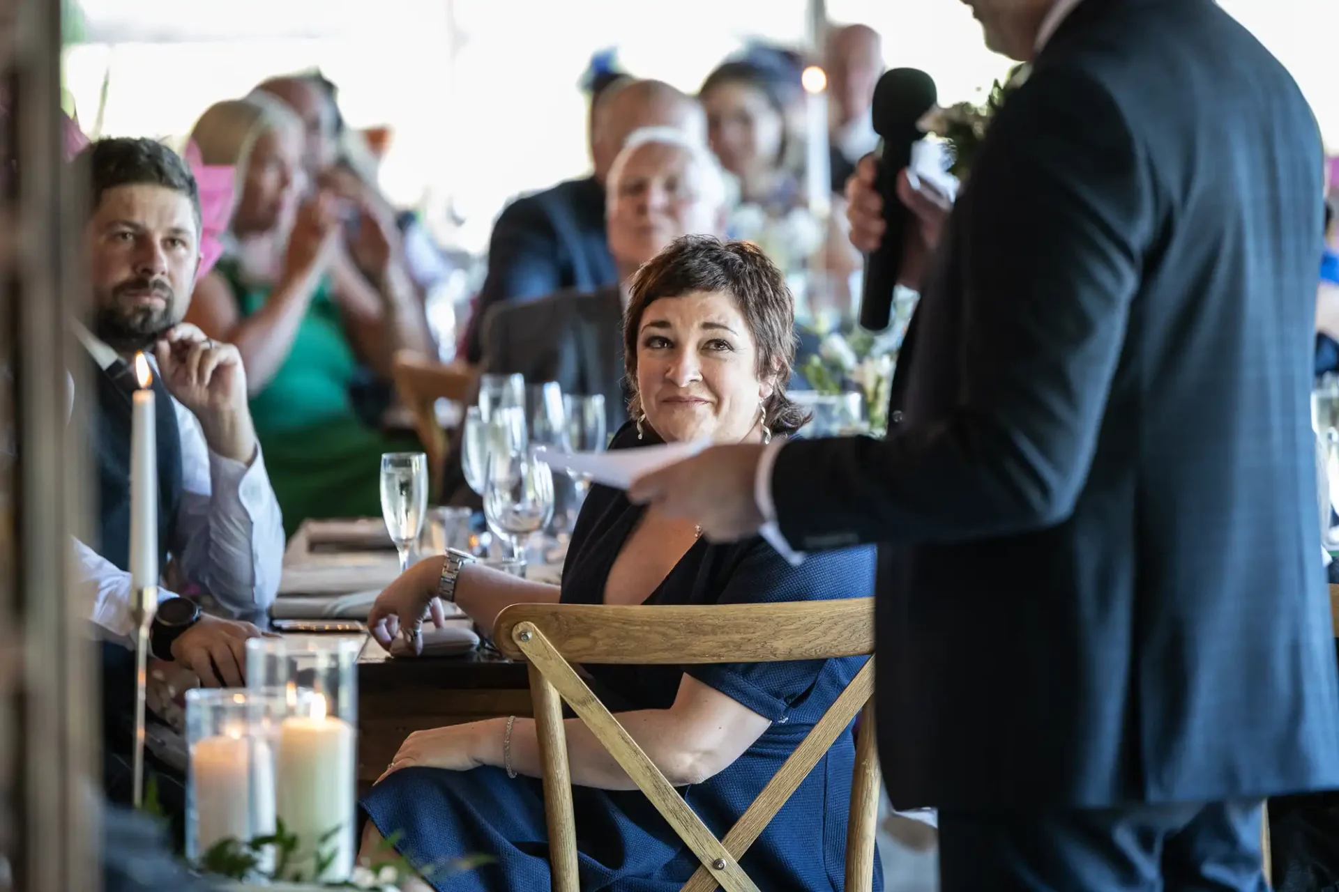 A woman seated at a table looks toward a speaker holding a microphone. The setting appears to be a formal gathering with attendees clapping.