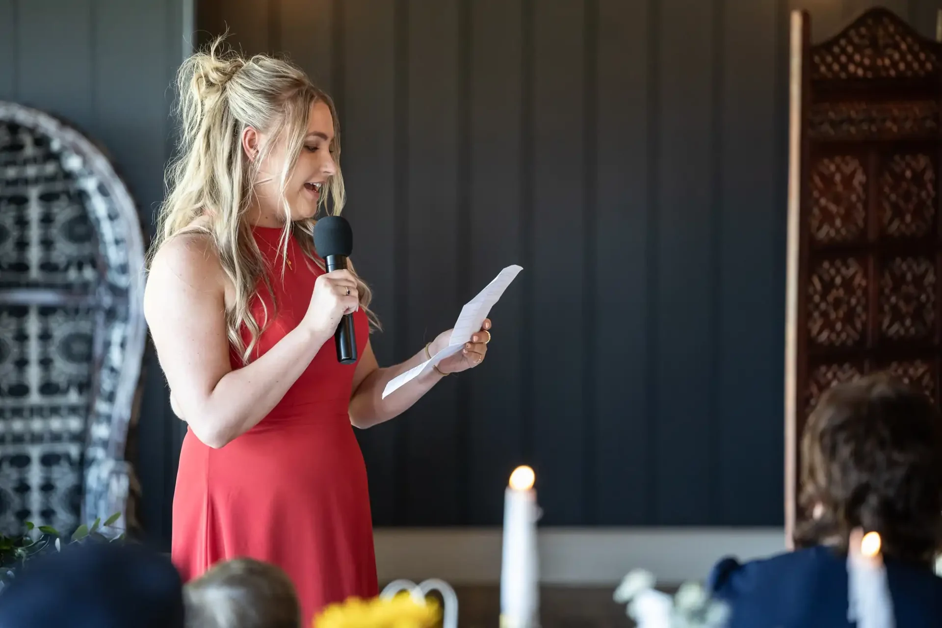 Woman in a red dress standing and speaking into a microphone while holding a piece of paper, with blurred people and candles in the foreground.