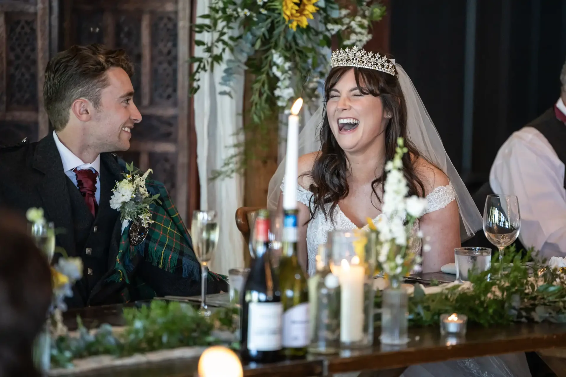 Bride and groom seated at a wedding table, adorned with candles and greenery. The bride is laughing while wearing a tiara and veil; the groom is smiling at her.