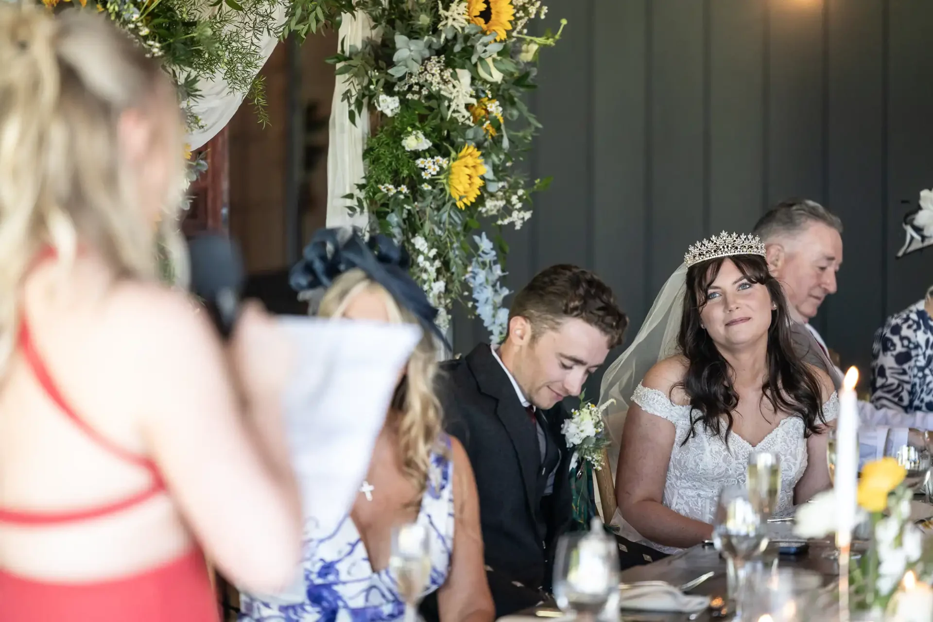 A bride listens during a wedding speech, seated beside the groom at a decorated table with flowers.