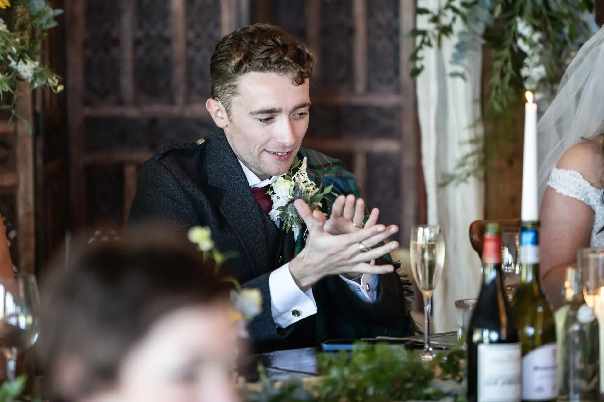 A man in a dark suit claps his hands, seated at a decorated table with a champagne glass and bottles.