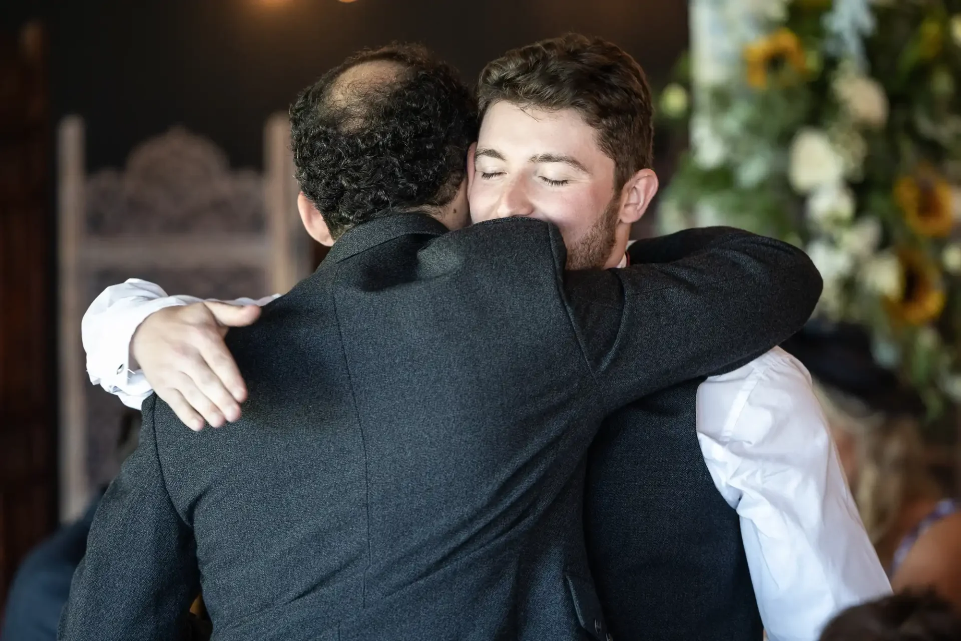 Two people warmly embrace at an indoor event, with one person facing the camera and the other facing away. They are dressed in formal attire.