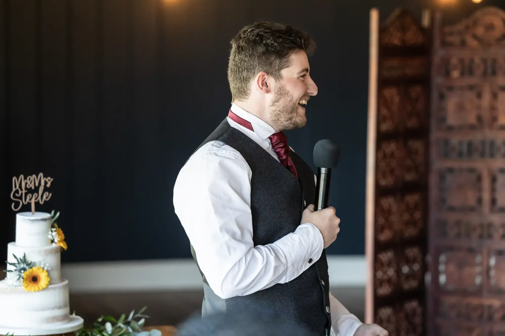 Man in a vest and tie holds a microphone, speaking and smiling, with a decorated cake on a table nearby.