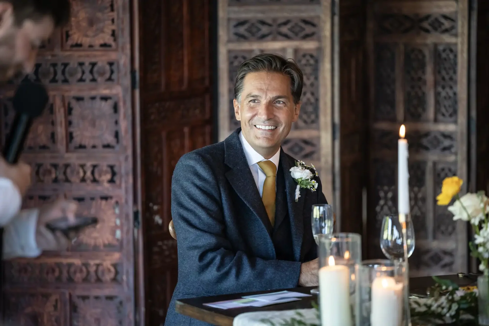 A person in a suit sits at a decorated table with candles and flowers, smiling, as another person speaks into a microphone off to the side. Ornate wooden panels are in the background.