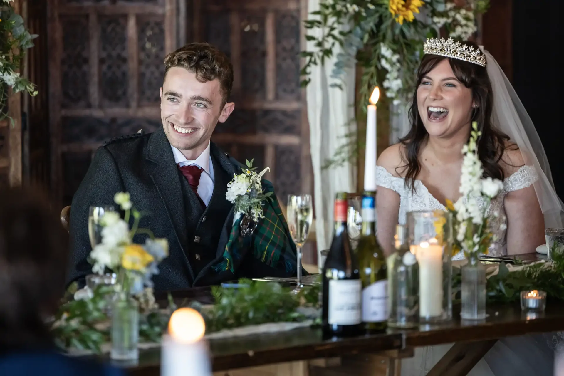 A bride and groom sit at a decorated table, smiling and laughing. The bride wears a tiara and off-shoulder dress, while the groom is in a suit with a tartan detail. Candles and flowers are on the table.