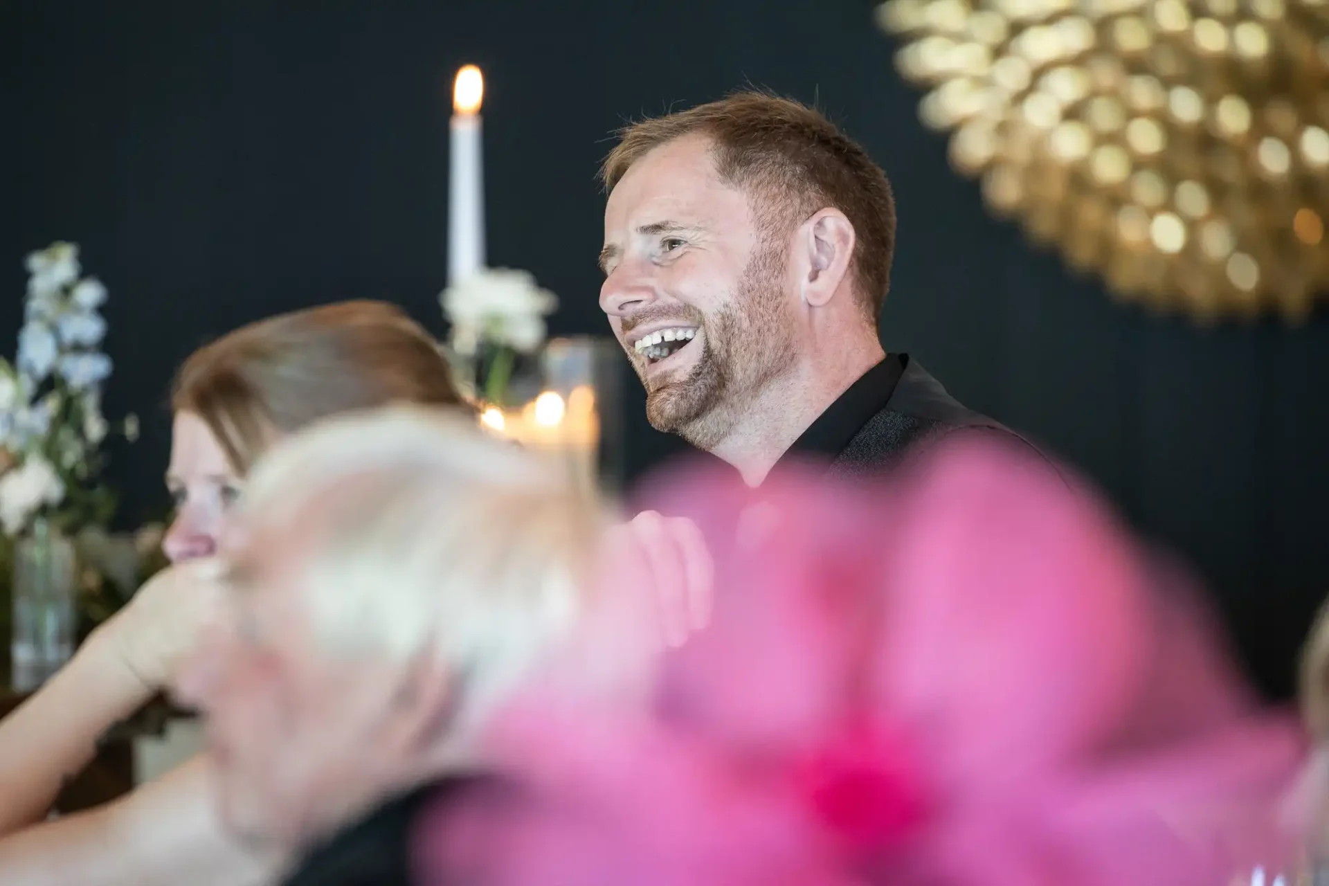 A man is laughing at a formal indoor event with candles and floral decorations in the background.