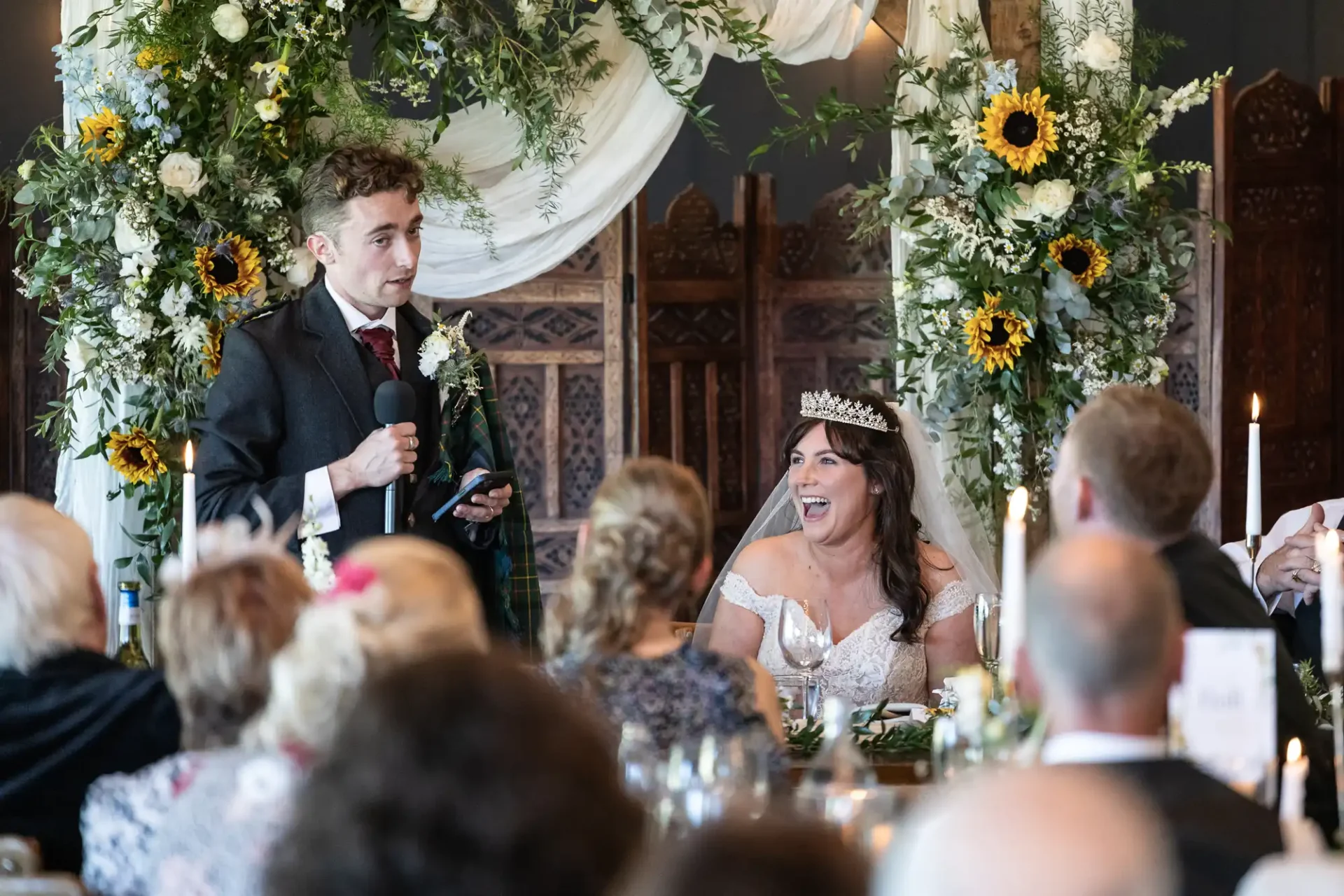 A groom gives a speech at a wedding reception. The bride, seated beside him, laughs. The room is decorated with floral arrangements and white drapery. Guests are seated at tables.