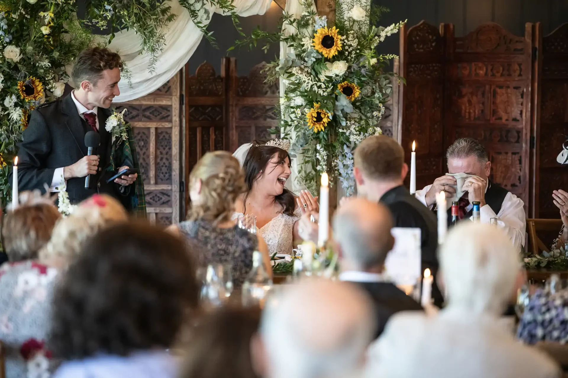 Wedding reception scene with a bride and groom at a table, surrounded by guests. A man in a suit is speaking, while another man wipes tears. Floral decorations and candles adorn the setting.