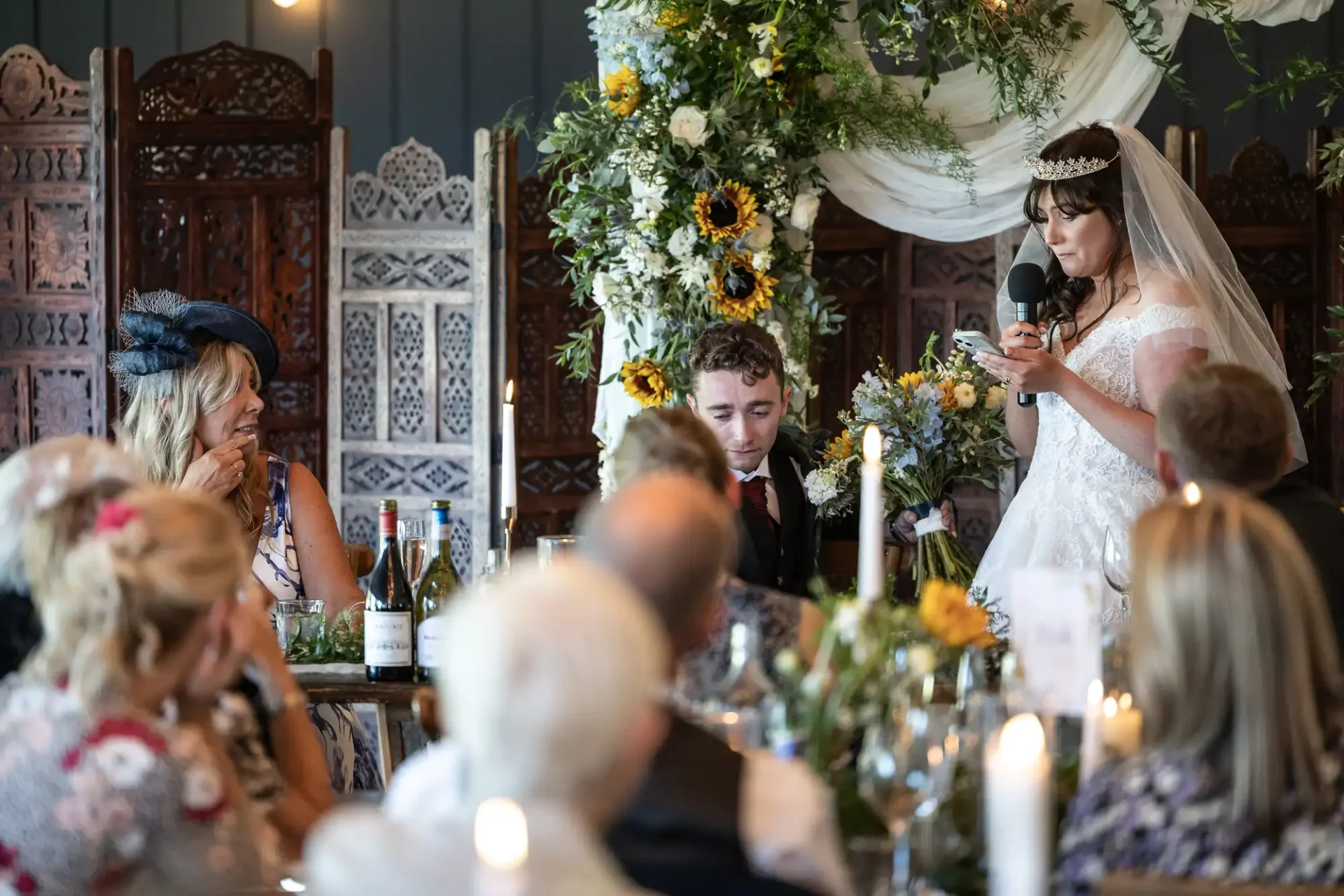 Bride giving a speech with a microphone at a decorated table during a wedding reception, surrounded by guests.