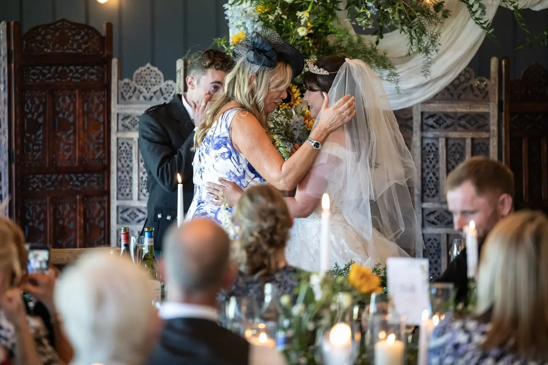 A bride in a white dress and veil embraces a woman in a floral dress at a wedding reception with guests seated and watching.