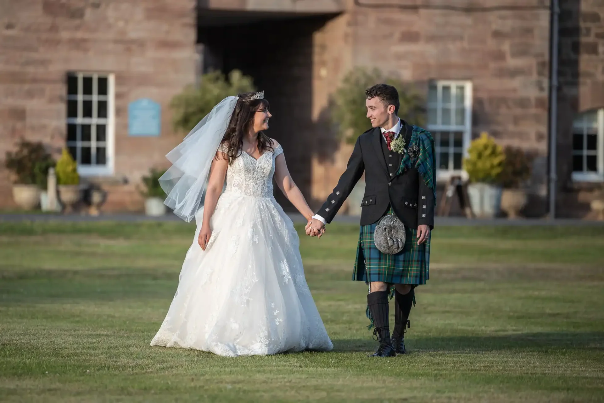 Bride in a white gown and groom in a kilt hold hands, walking on grass in front of a stone building.