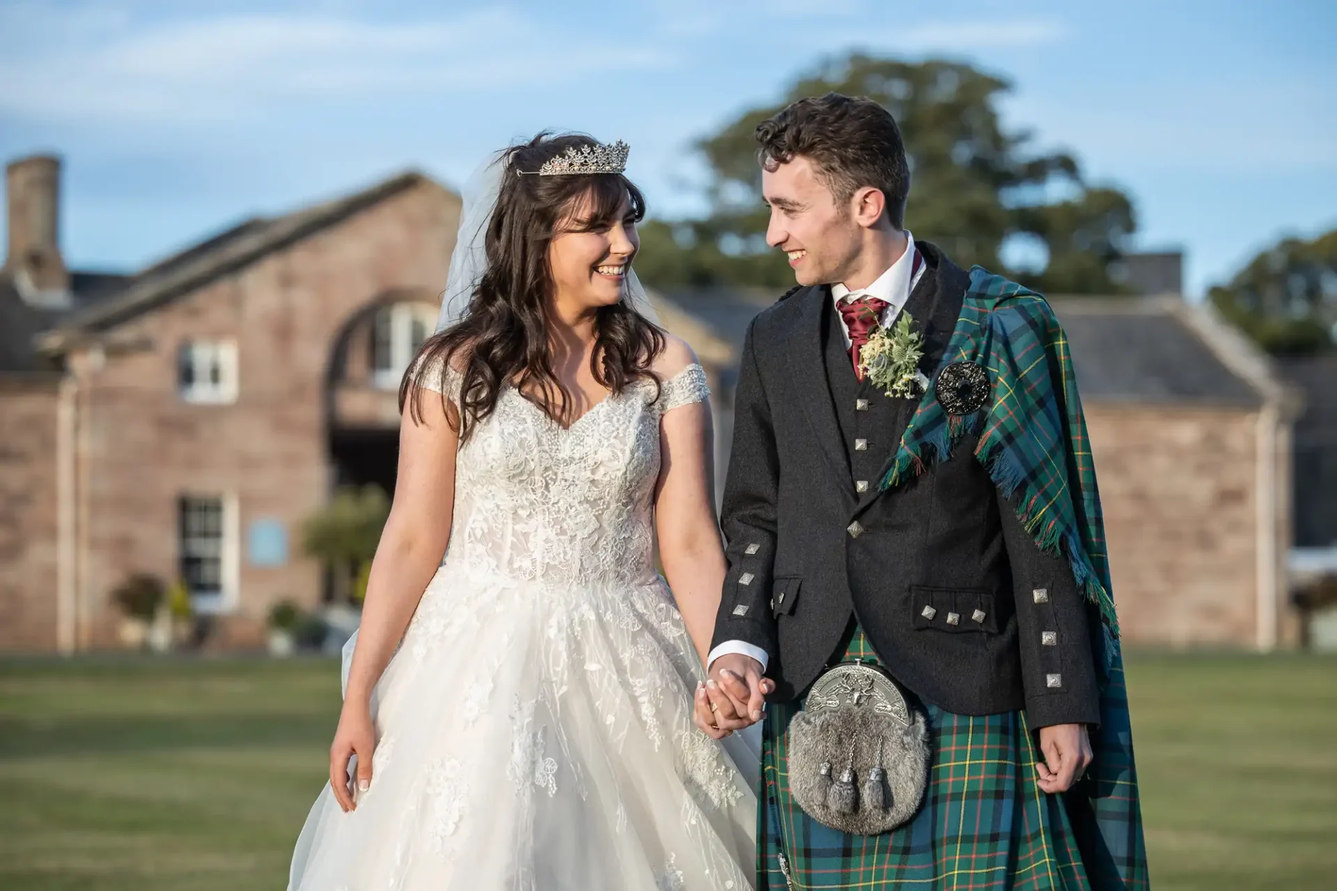 A bride in a white gown and tiara holds hands with a groom in a green tartan kilt and dark jacket. They walk happily on a grassy area in front of a stone building.