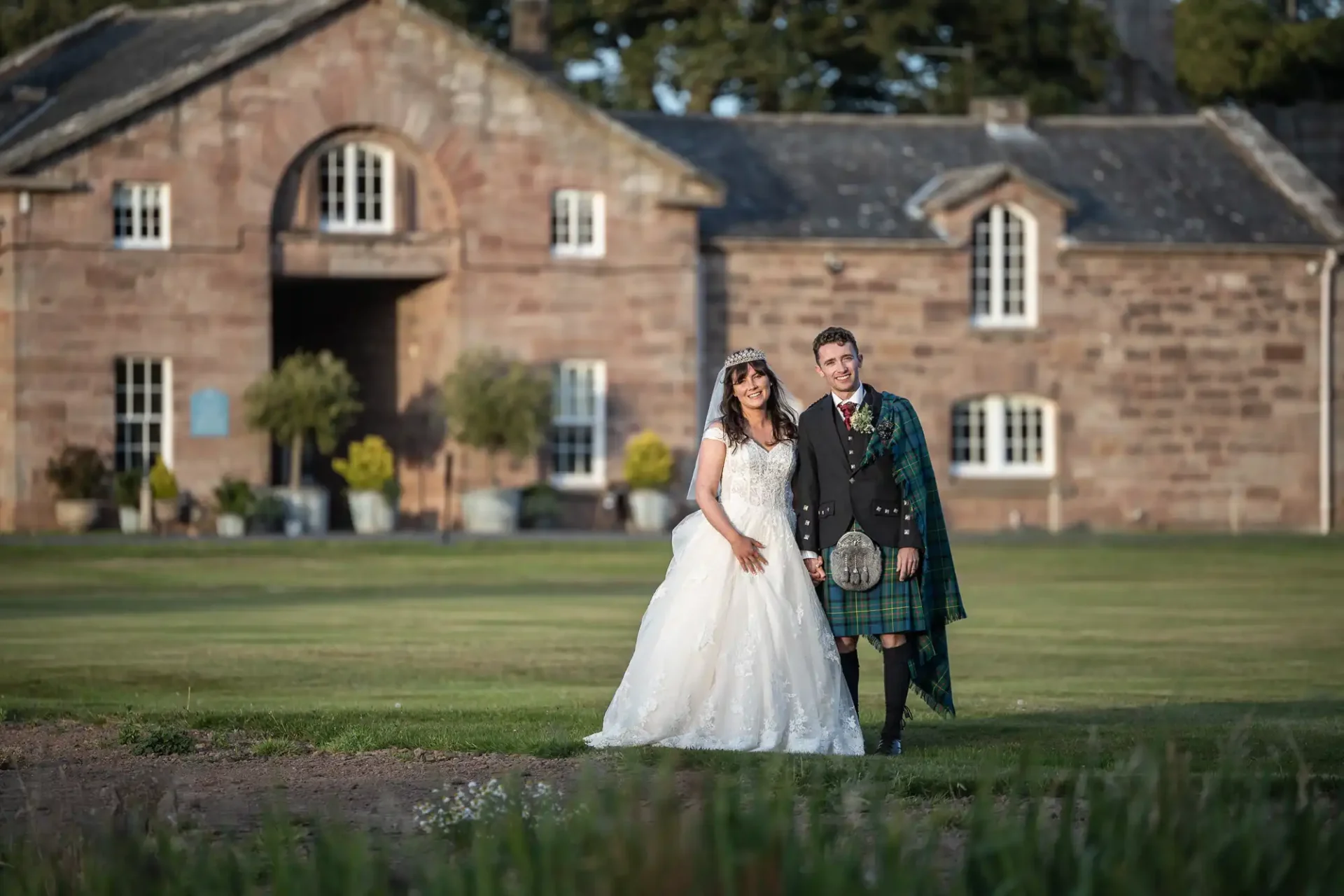 A couple stands in wedding attire, with the bride in a white gown and the groom in a traditional Scottish kilt, in front of a historic stone building.