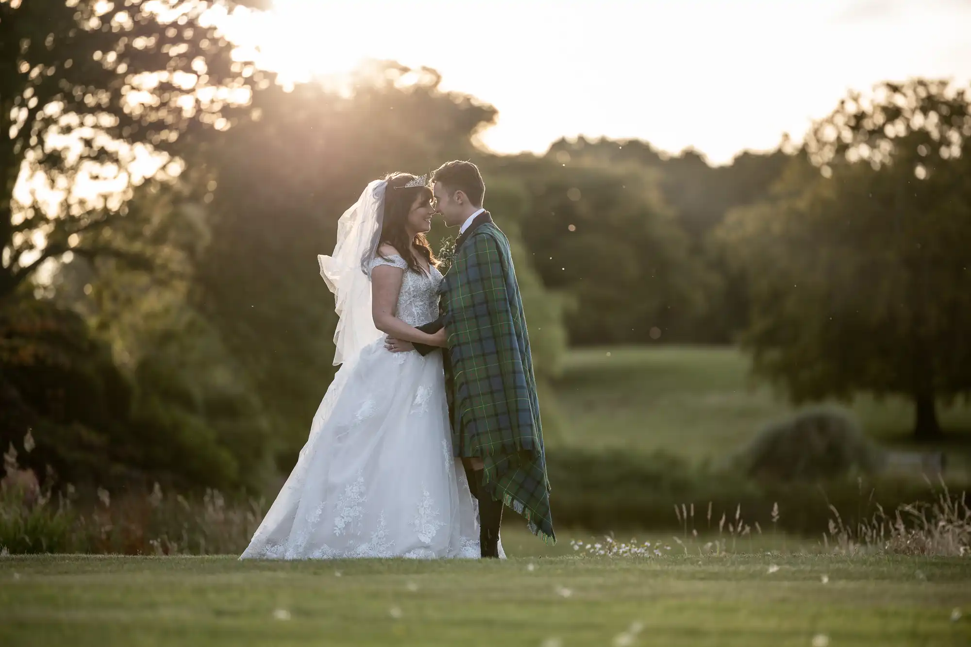 wedding photographers at Dunglass Estate: Bride and groom standing together in a grassy field, with the groom wearing a tartan shawl. Sunlight filters through trees in the background.