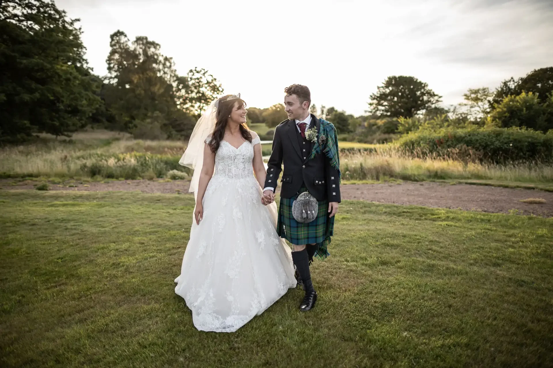 A couple in wedding attire holds hands while walking on grass. The groom wears a kilt, and the bride is in a white gown. Trees and sky are in the background.