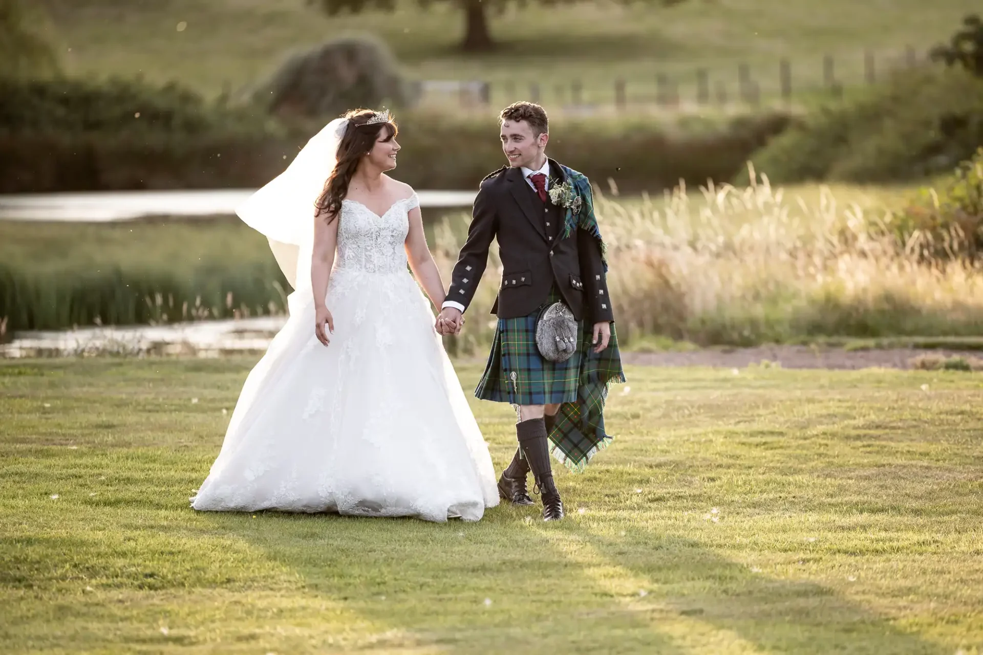 A bride and groom holding hands, walking on a grassy field; the groom is wearing a kilt and the bride has a veil.