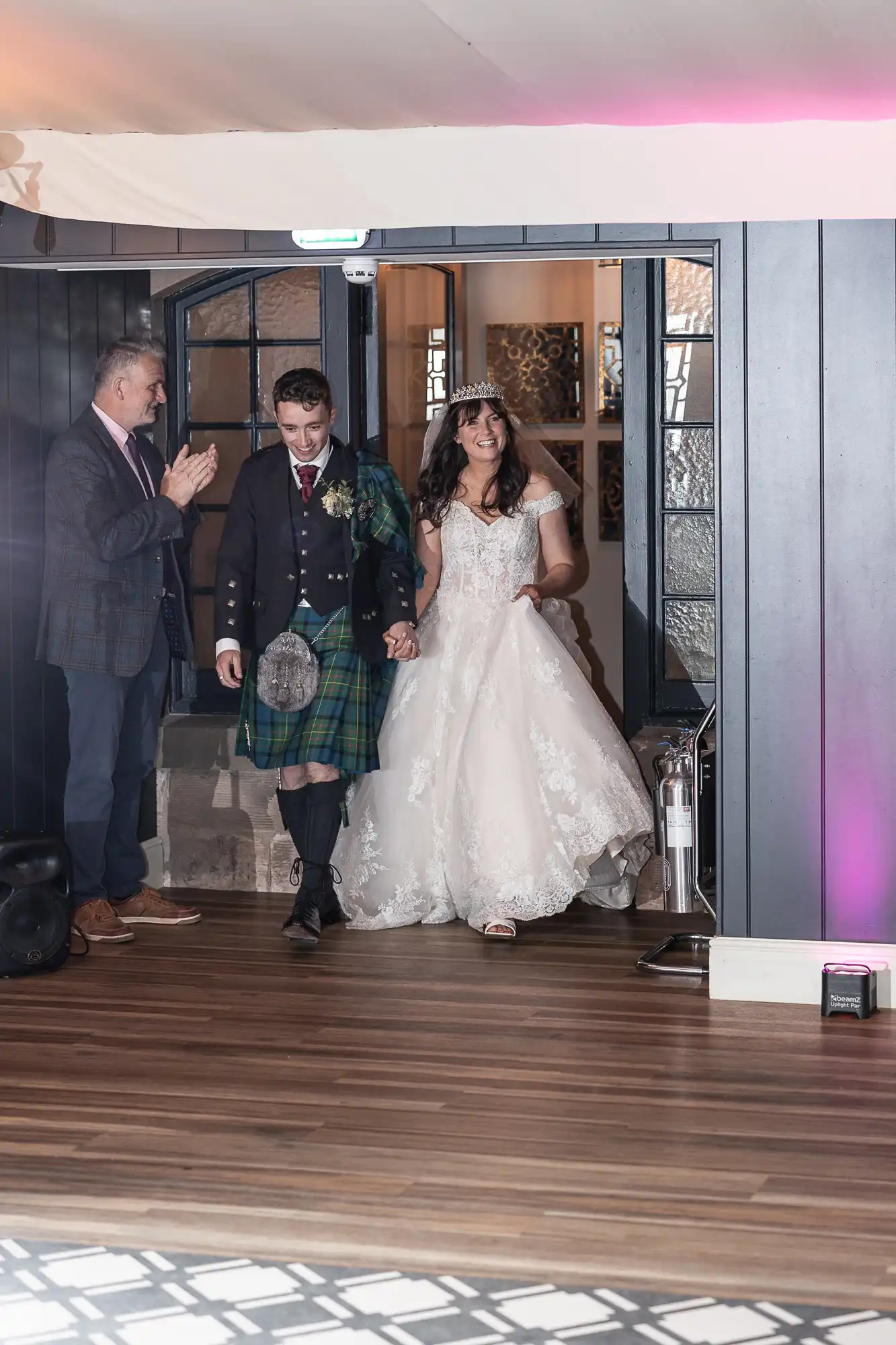 Bride and groom in formal attire, including a tartan kilt, entering a room with a man clapping nearby.