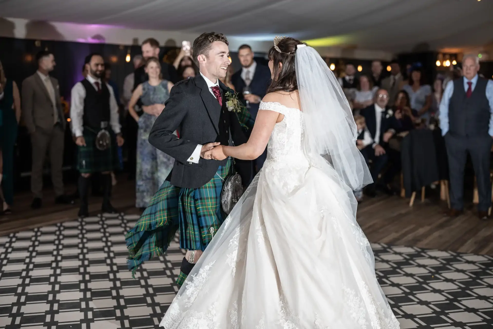 A couple in wedding attire dances on a checkered floor, surrounded by people. The groom wears a kilt and the bride wears a white gown with a veil.