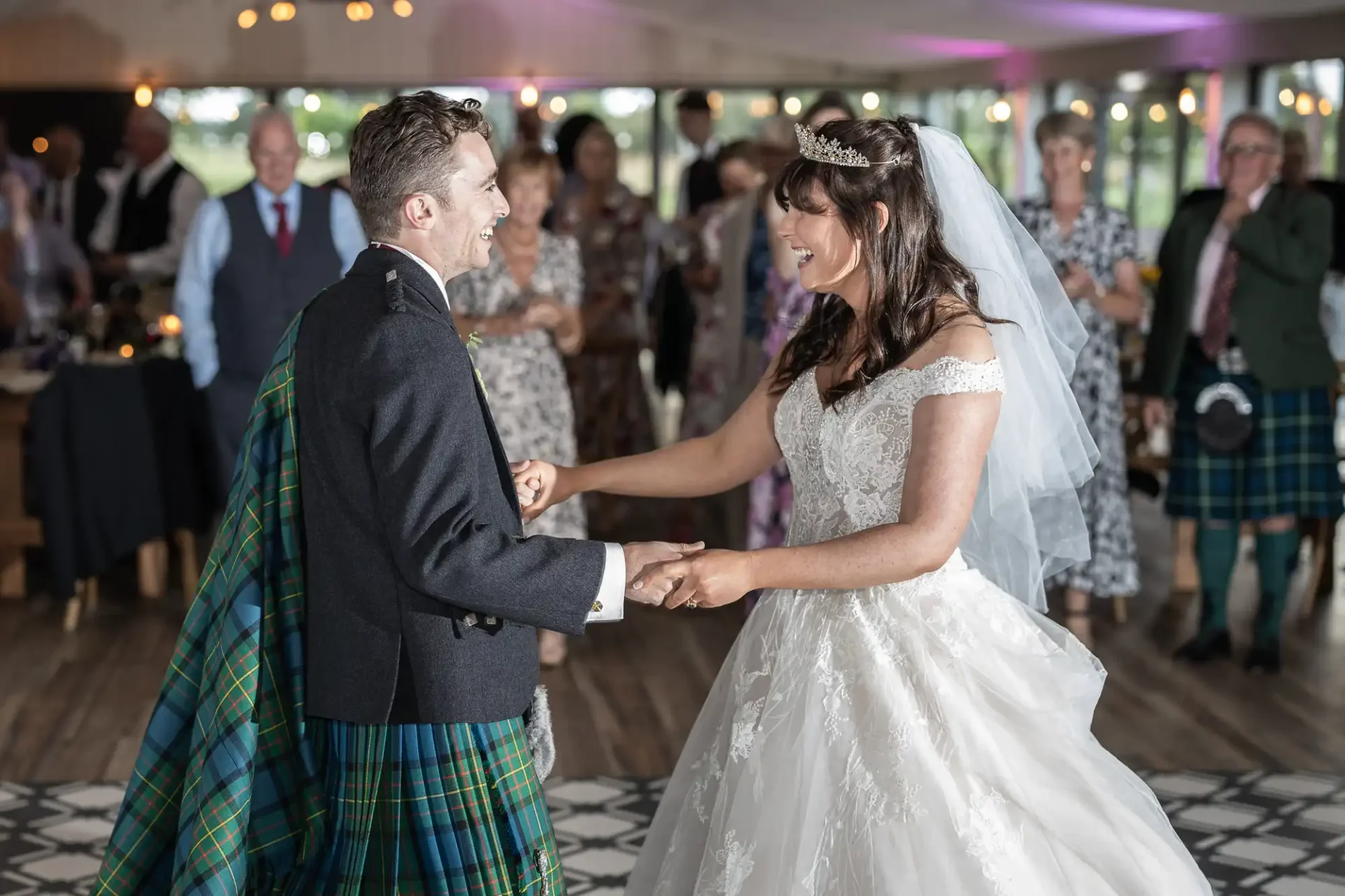 A bride and groom dance together at their wedding, surrounded by guests. The groom wears a kilt, and the bride wears a white gown and veil.