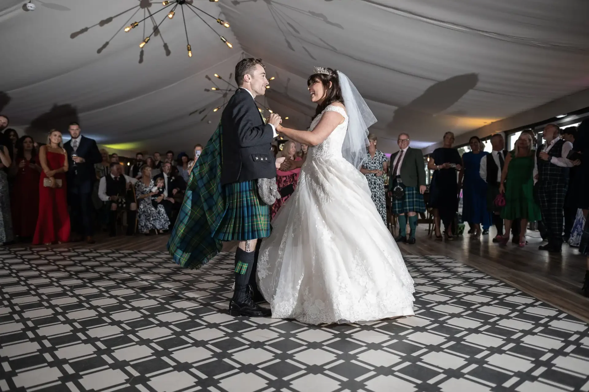 Bride and groom dance at a wedding reception. The groom wears a kilt, and the bride is in a white gown. Guests watch them under a tent with string lights.