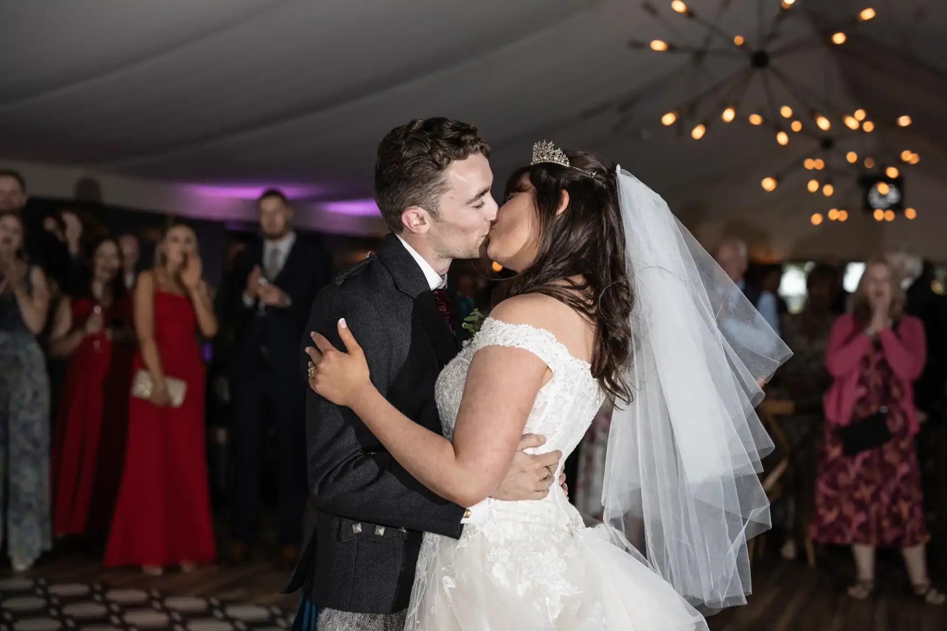 Bride and groom kissing on the dance floor during their wedding reception, surrounded by guests inside a decorated venue.