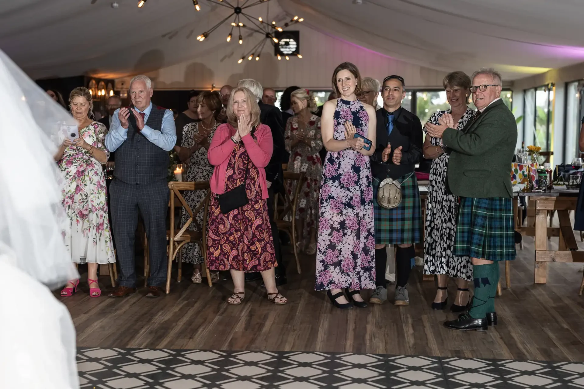 Guests in floral and tartan attire applaud at a wedding reception in a decorated hall.