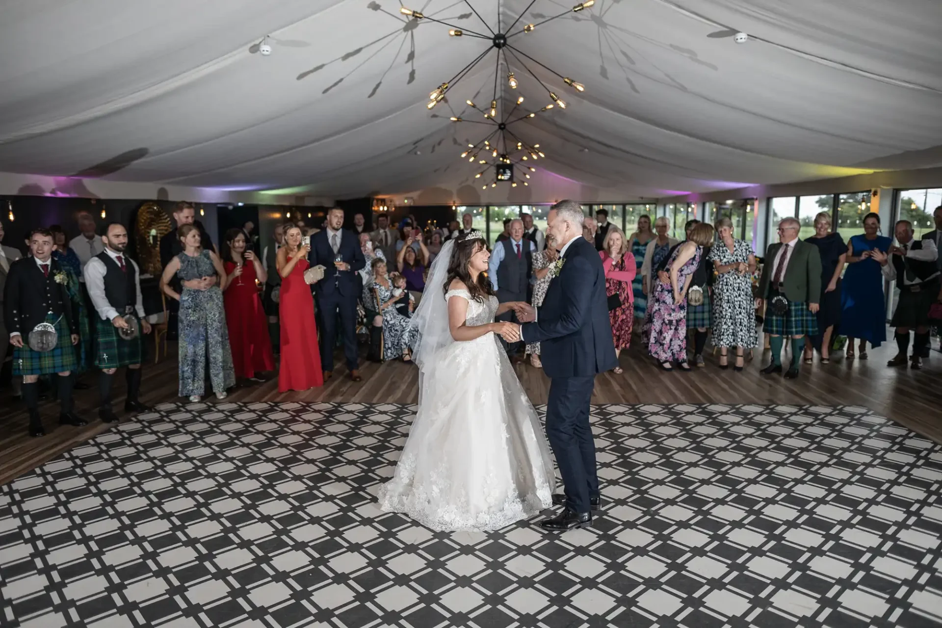 A bride and groom dance in a tented venue with a patterned floor, surrounded by guests. Some attendees wear Scottish kilts. The ceiling features decorative lighting.