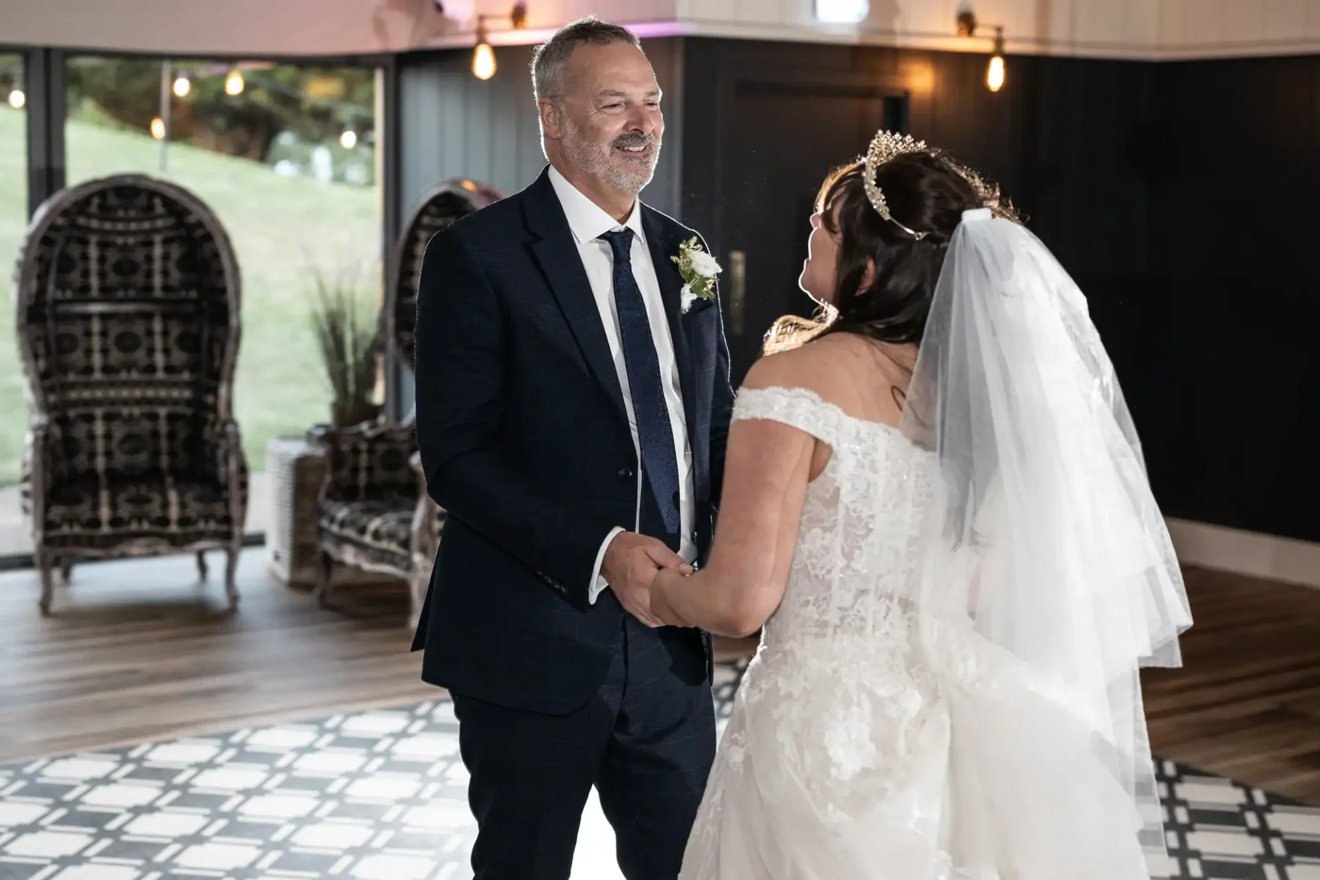 A couple dances in a room with patterned flooring and black walls. The man wears a dark suit, and the woman wears a white wedding dress and veil under soft lighting.