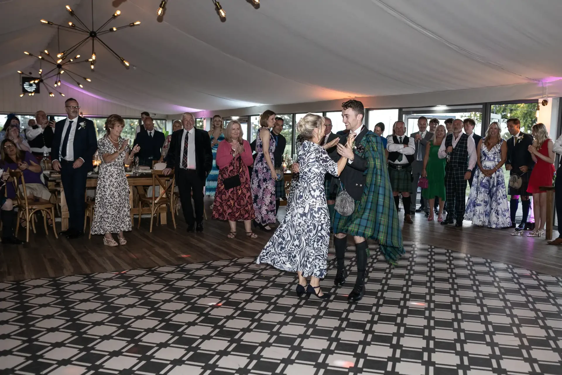 A couple dances on a patterned floor at an indoor event. Onlookers, some in formal and kilts attire, watch the dance under decorative ceiling lights.