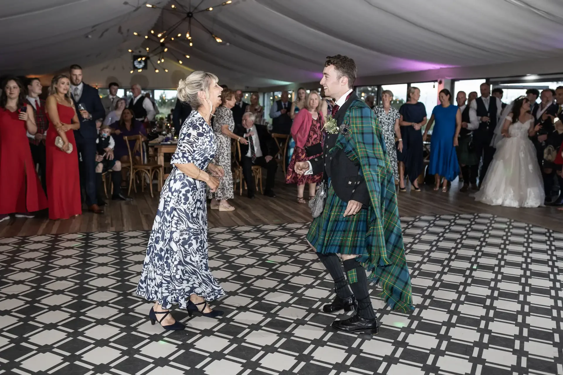 A man in a tartan outfit dances with a woman in a patterned dress on a checkered floor at a formal event with guests watching.