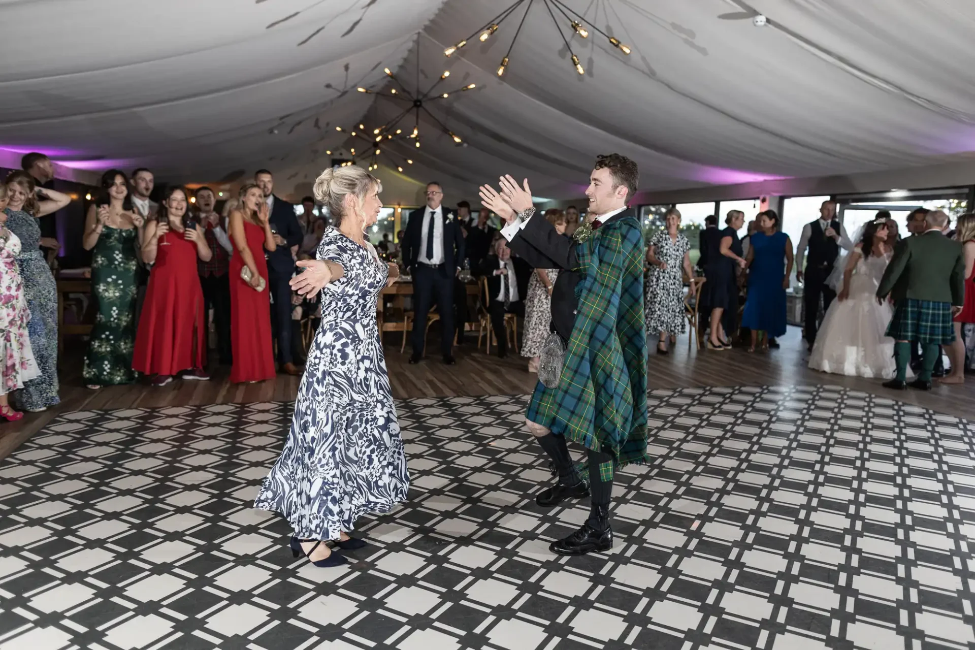 A man in a plaid kilt and a woman in a patterned dress dance on a checkered floor at an event. People watch in the background under a tent with string lights.