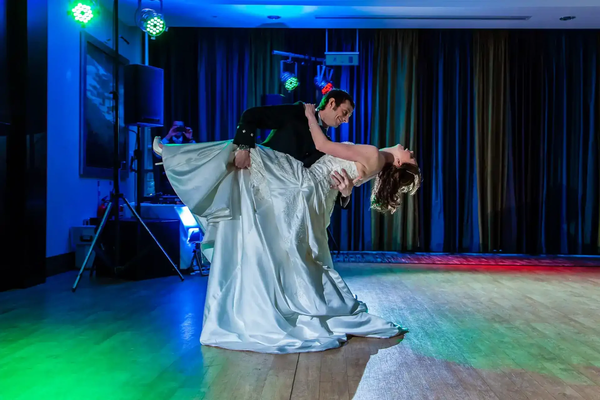 A bride and groom perform a dramatic dance dip on a wooden floor under colourful stage lights, with a photographer capturing the moment in the background at Norton House Hotel Garden Suite, Edinburgh.