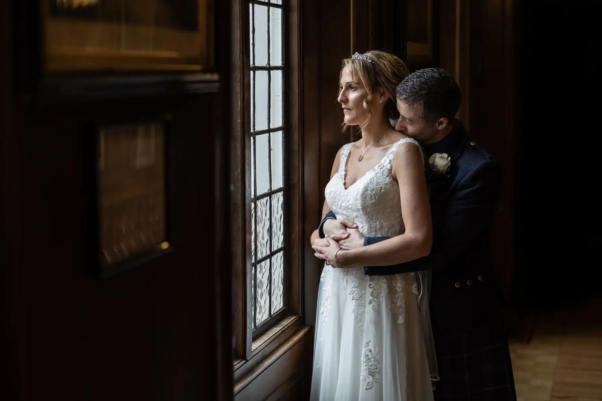 A bride in a white dress and a groom embrace by a window, looking outside in a dimly lit room with dark wooden walls at the Royal College of Physicians of Edinburgh.