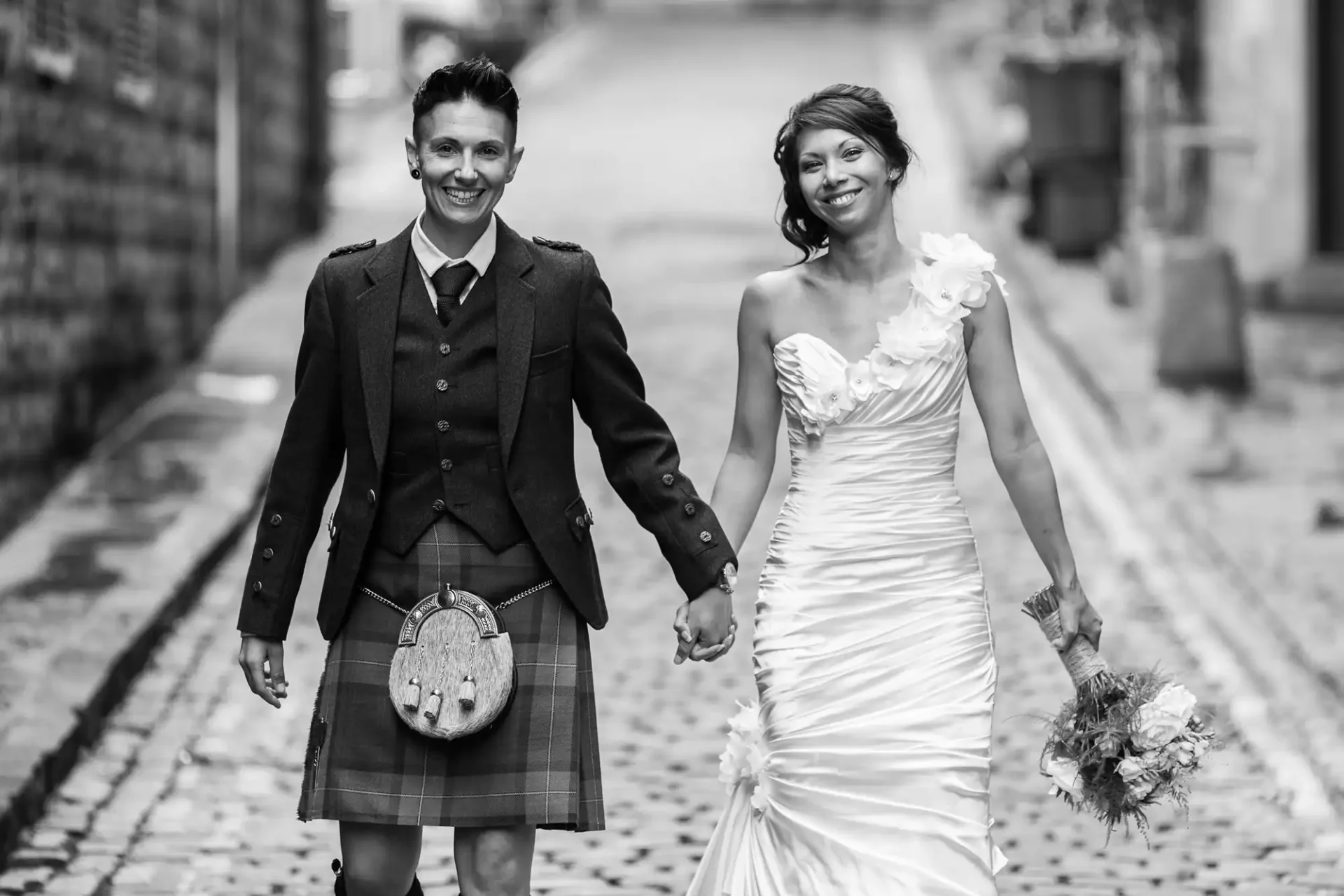 Two people are holding hands and smiling, one wearing a kilt and jacket, the other in a wedding dress, standing on a cobblestone street at The Rowntree, Edinburgh.