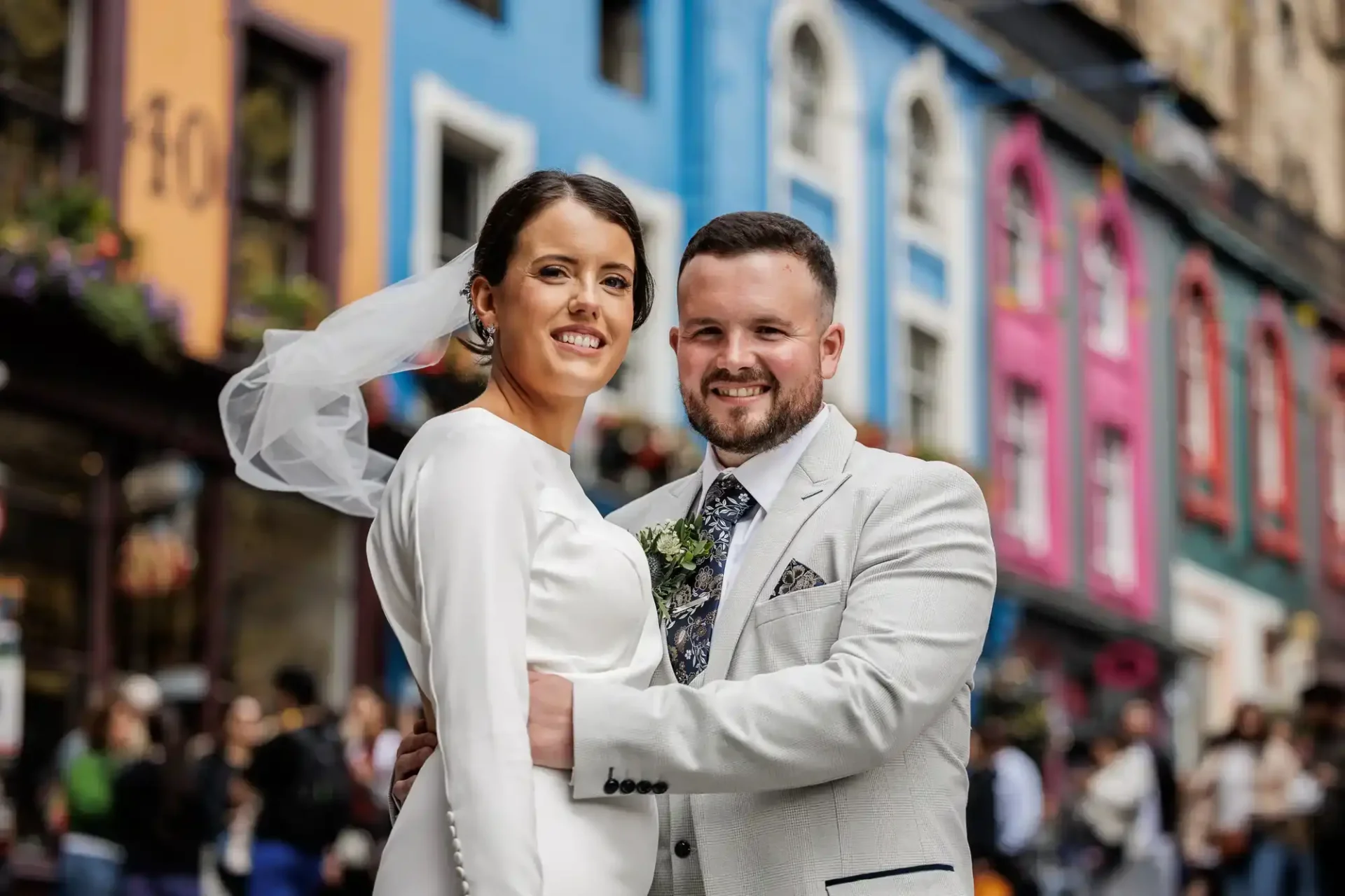 A bride and groom in wedding attire pose together on a city street with colourful buildings in the background at Victoria Street, Edinburgh.