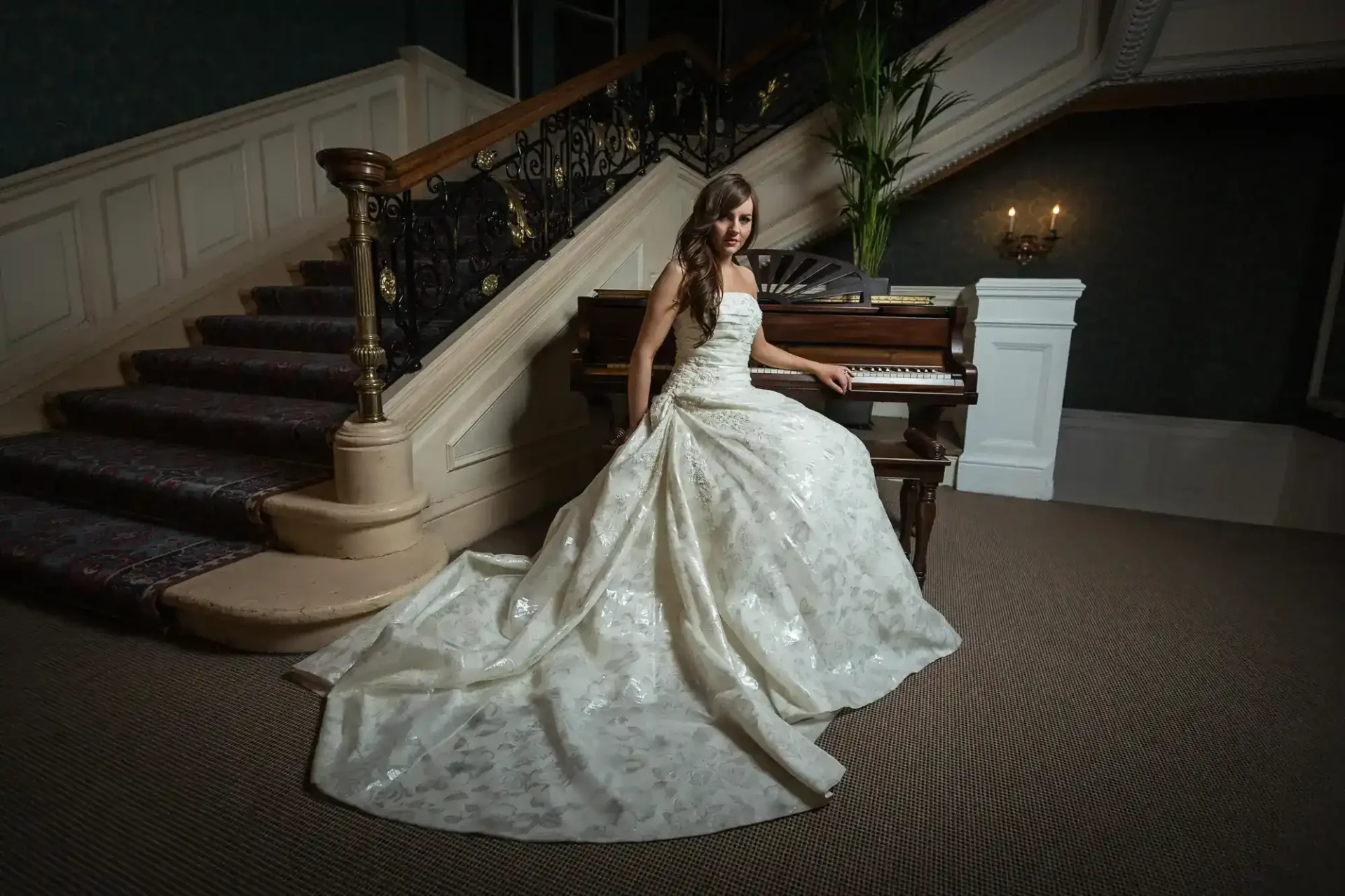 A woman in a white wedding dress sits at a piano in an elegant room with a carpeted staircase and ornate railing in the background at the Balmoral Hotel, Edinburgh.
