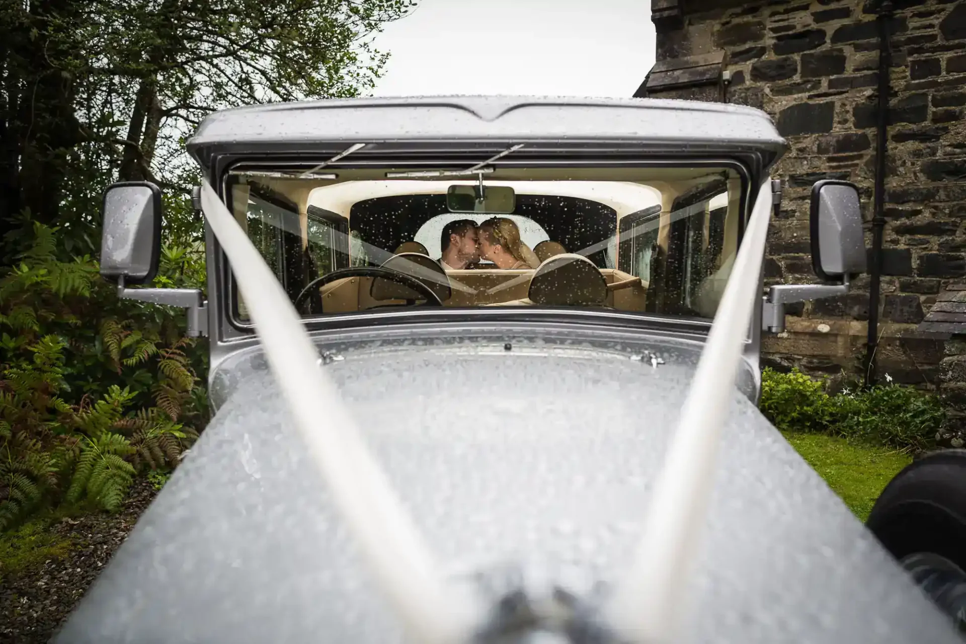 A couple sits inside a vintage car on a rainy day, seen through the windshield with raindrops and wedding ribbons visible on the hood at Mansfield Traquair, Edinburgh.