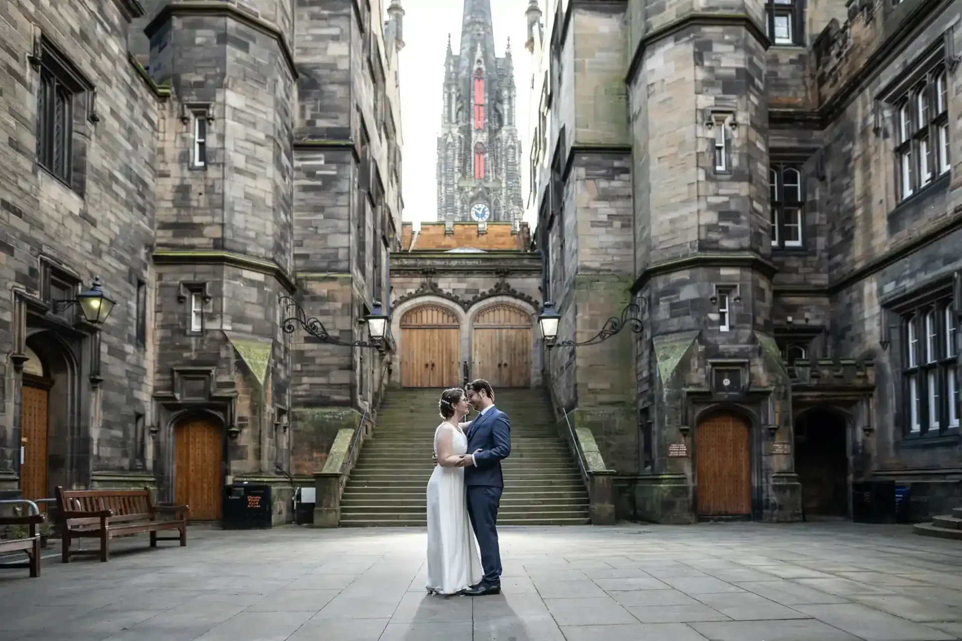 A couple in wedding attire stands together in a stone courtyard between historic buildings, with a tall gothic tower visible in the background at New College Quad, Edinburgh.