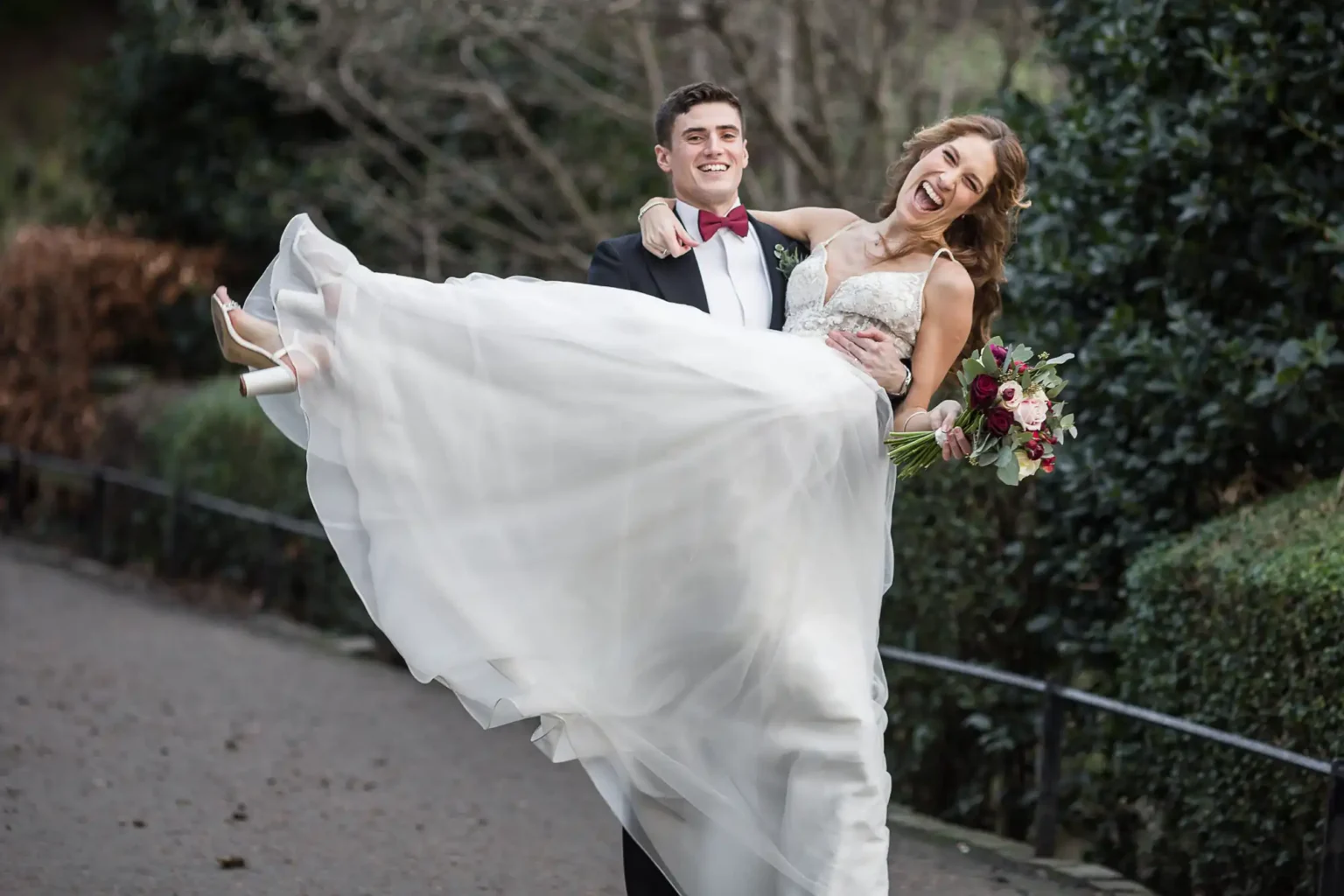 A groom in a black suit holds a smiling bride in a white wedding dress and holding a bouquet, outdoors on a pathway at Princes Street Gardens, Edinburgh.