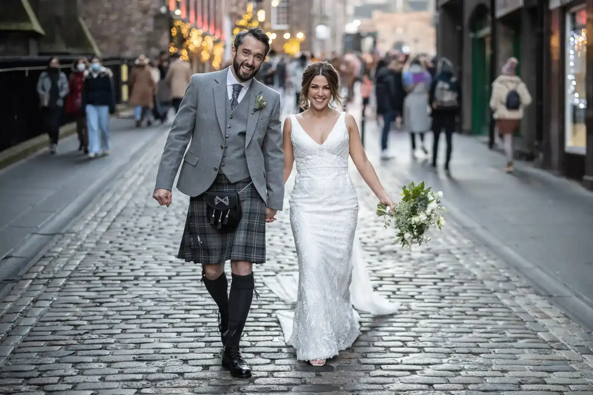 A bride in a white dress and a groom in a traditional Scottish kilt walk hand-in-hand down a cobblestone street, smiling, with people in the background at the Royal Mile, Edinburgh.