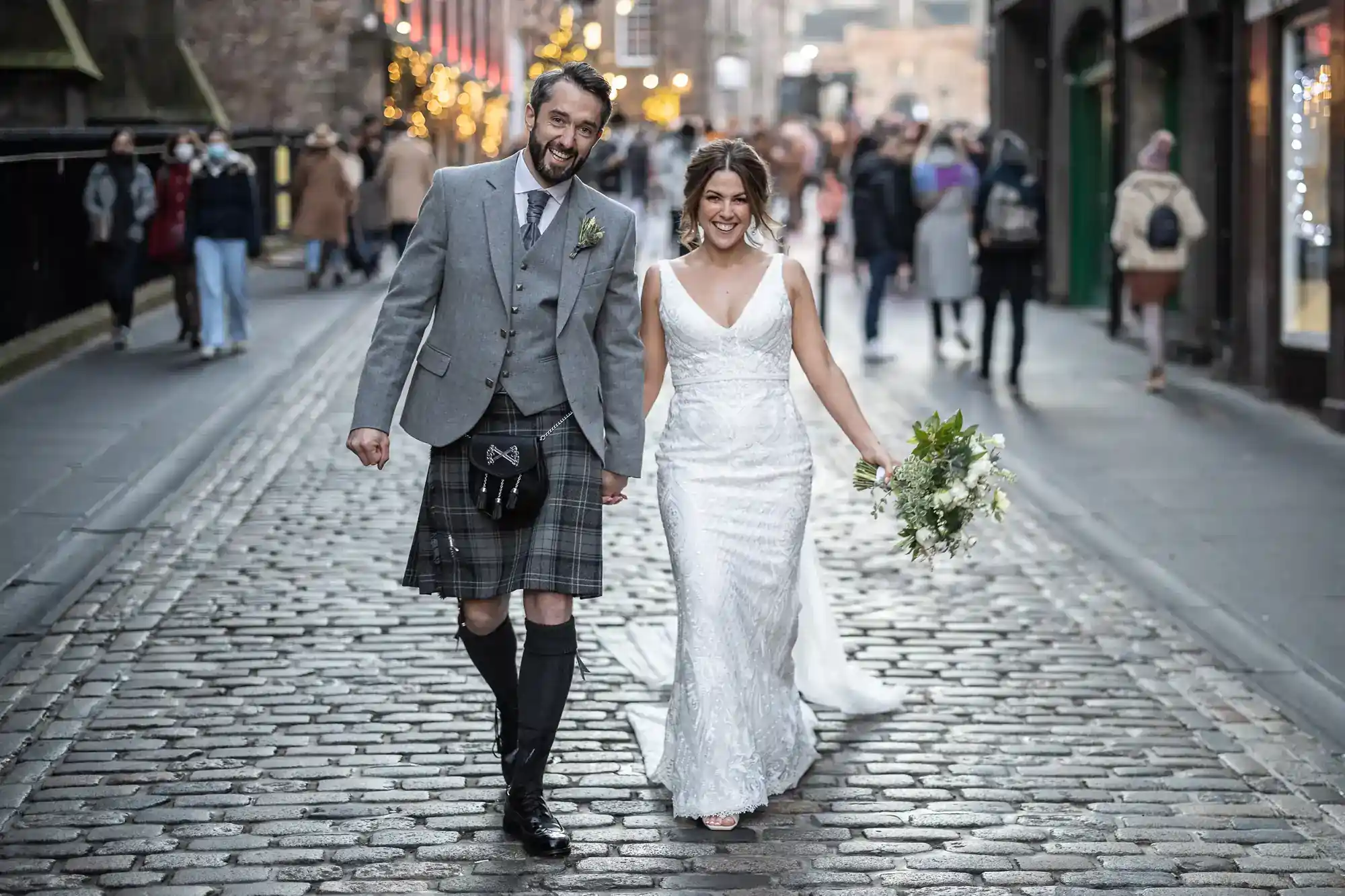 A bride in a white dress and a groom in a traditional Scottish kilt walk hand-in-hand down a cobblestone street, smiling, with people in the background at the Royal Mile, Edinburgh.