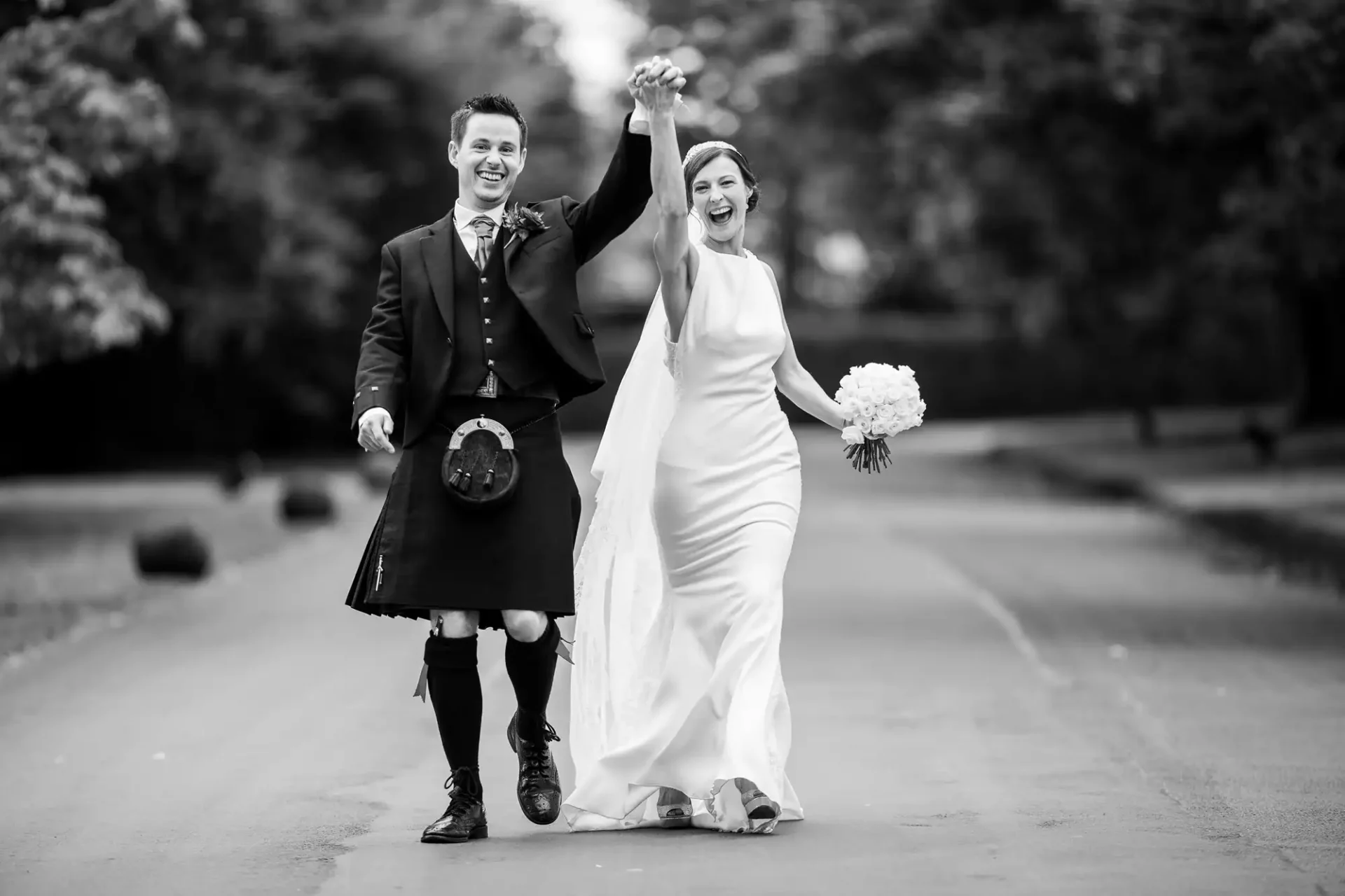 A smiling bride and groom walk hand in hand down an empty road; the groom wears a kilt, and the bride holds a bouquet and wears a long dress and veil at Prestonfield House, Edinburgh.