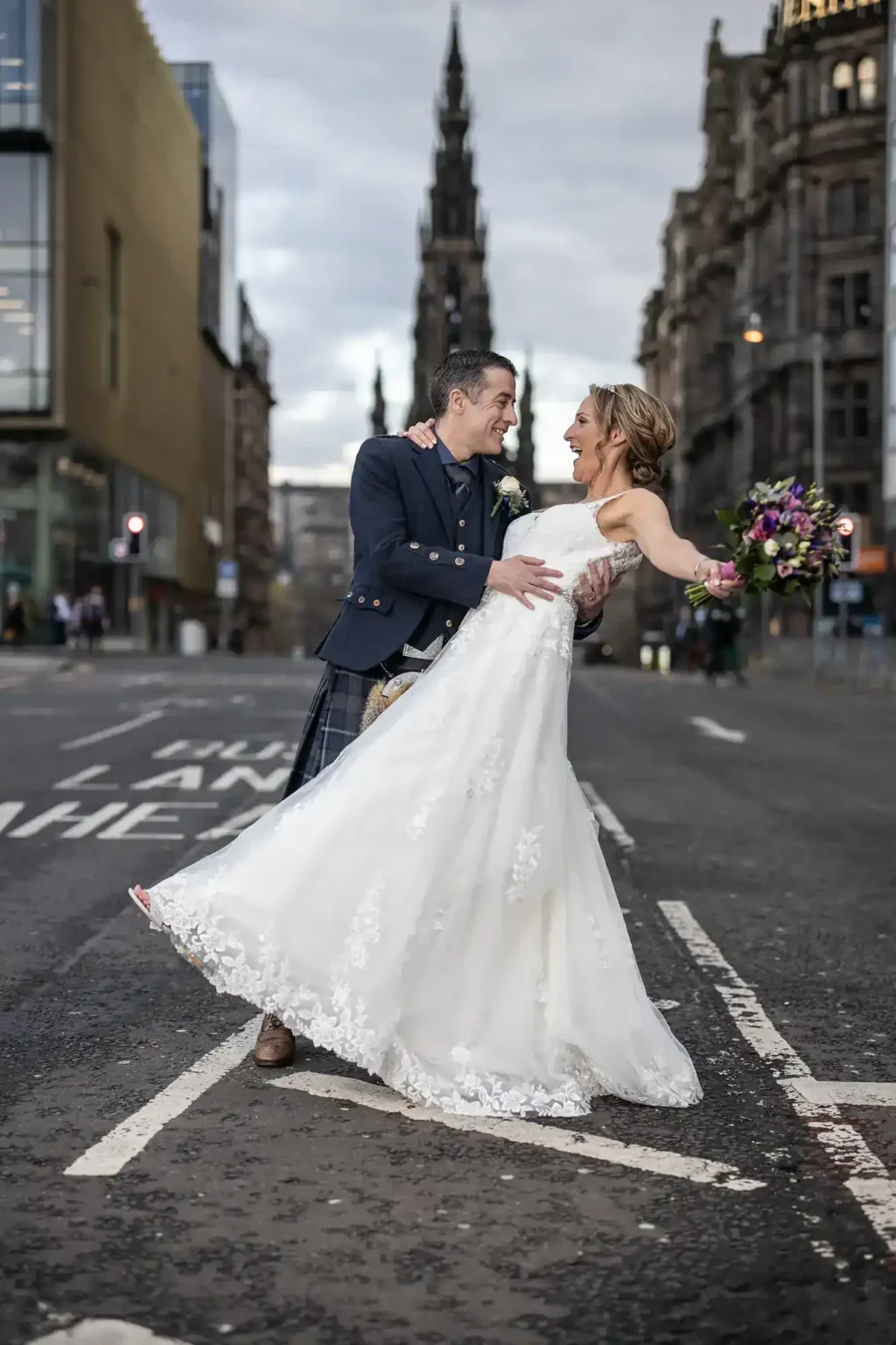 A bride and groom stand joyfully in the middle of a city street, with historic buildings and Scott Monument visible in the background at St Andrew Square, Edinburgh.