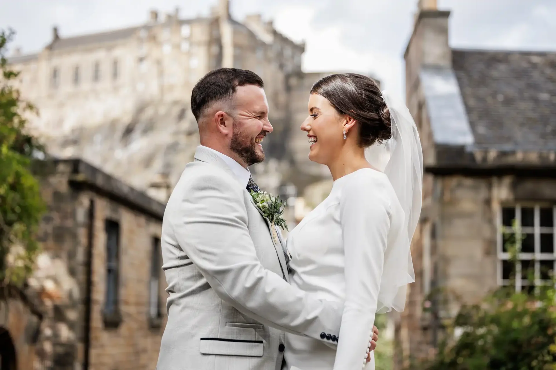 A bride and groom in white attire stand facing each other and smiling, with historic stone buildings and Edinburgh castle in the background, at The Vennel, Edinburgh.