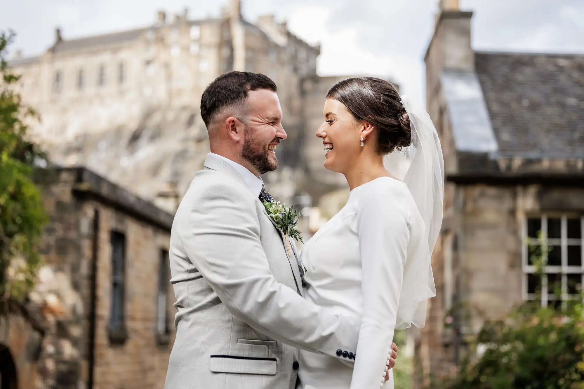 A bride and groom in white attire stand facing each other and smiling, with historic stone buildings and Edinburgh castle in the background, at The Vennel, Edinburgh.