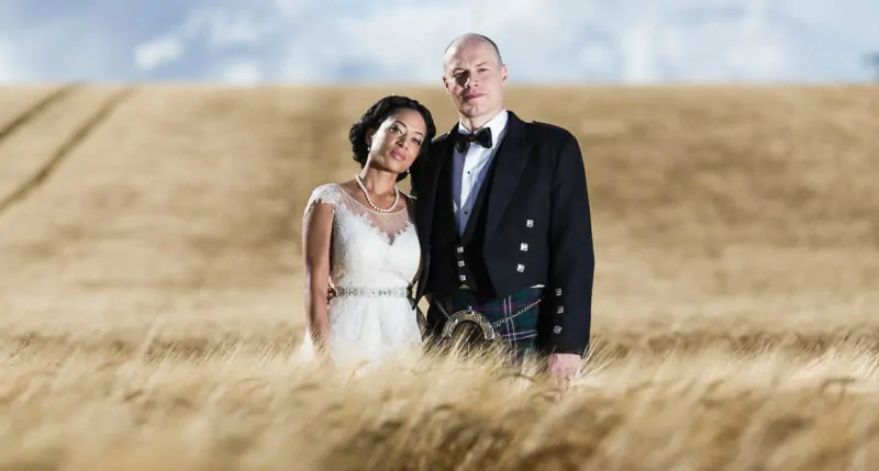 A bride and groom standing in a golden wheat field; the groom wears a kilt and the bride is in a white dress.