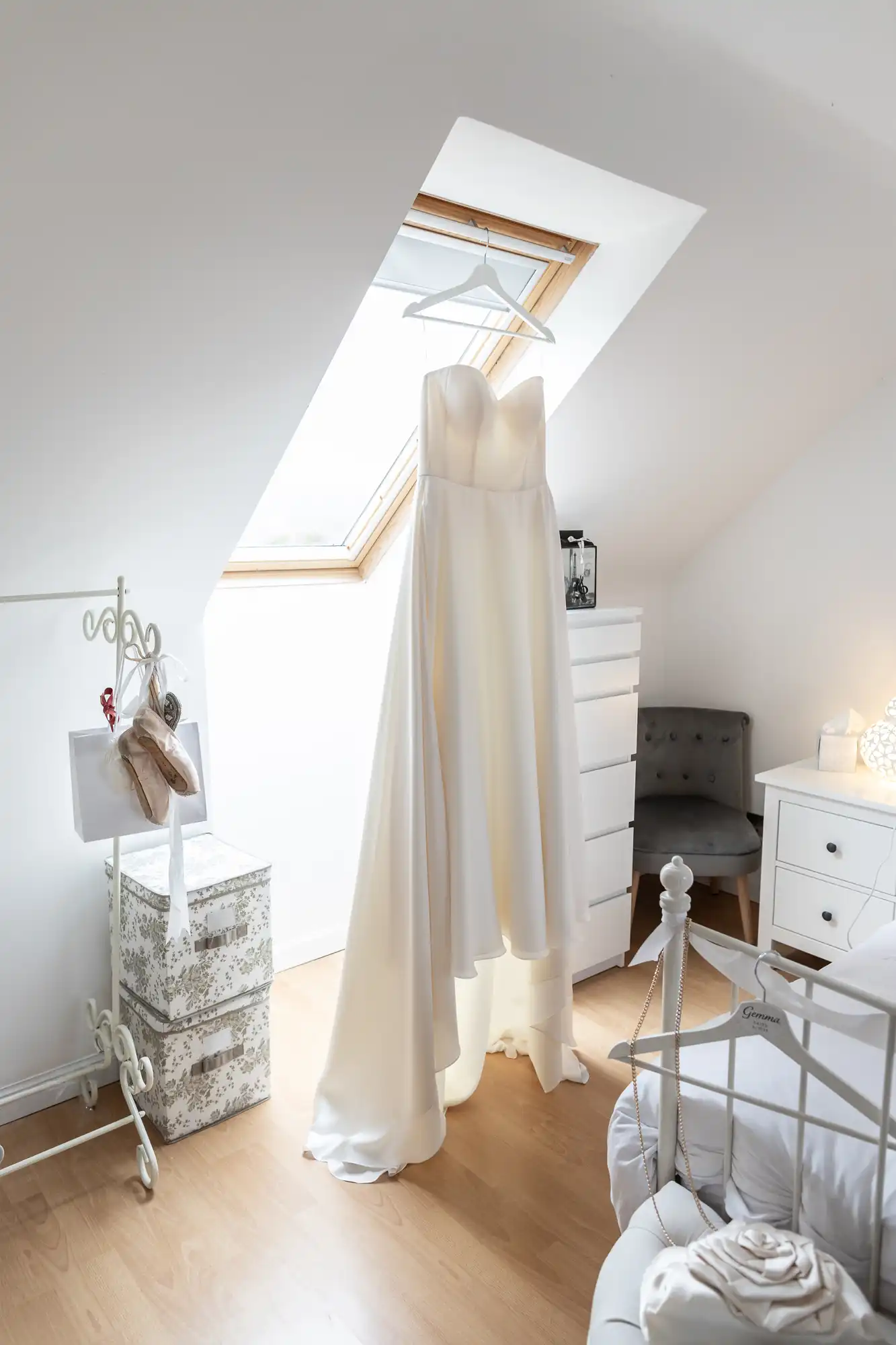 A white wedding dress hangs in a bright room with a skylight, near a gray upholstered chair and decorative storage boxes.