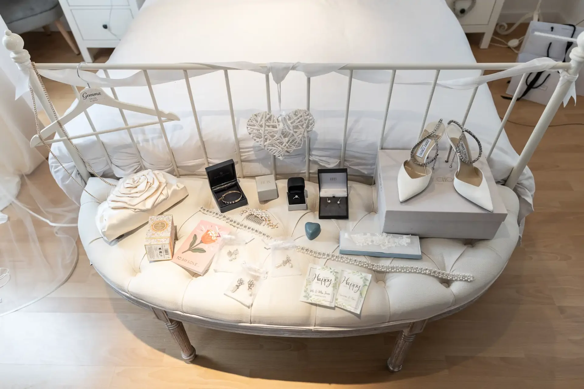 White bench with wedding items: shoes, jewelry boxes, flower, hanger, and cards. Bed and nightstand in the background.