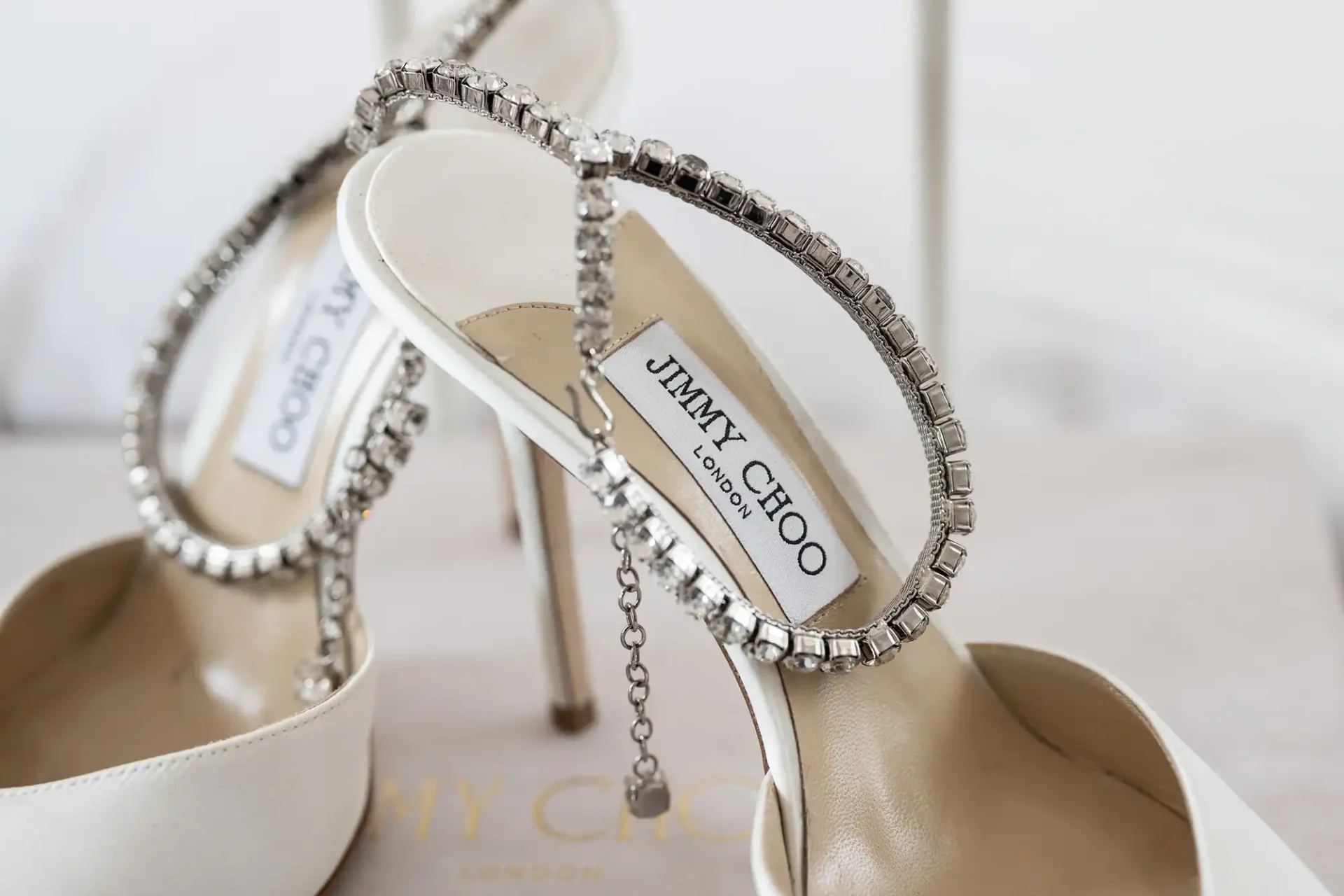 A pair of white high-heeled shoes with rhinestone straps and a label reading "Jimmy Choo London" on the insole.