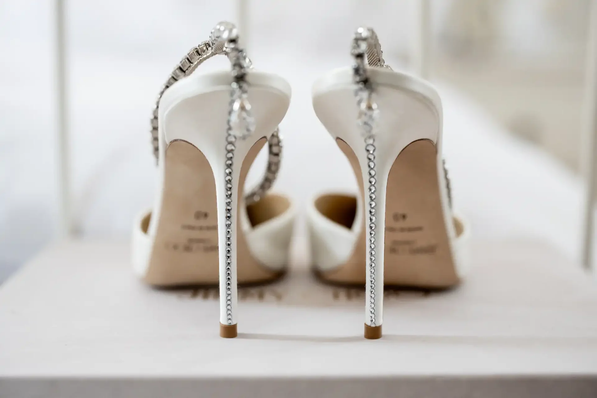 The image shows a pair of elegant white high-heeled shoes with jeweled straps, viewed from the back, resting on a surface.